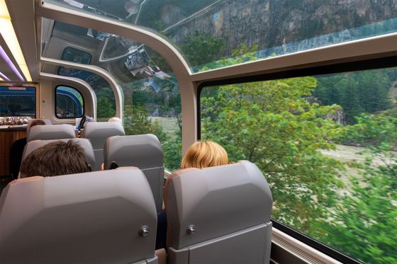 Passengers in a train with large windows enjoy scenic views of lush greenery and mountains through the glass roof and side.