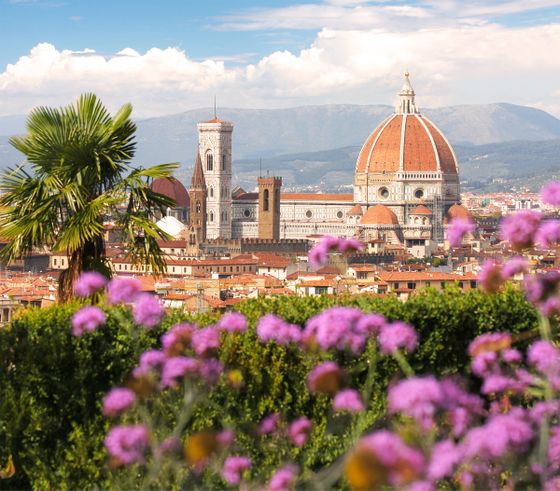 Florence Cathedral with its iconic dome and bell tower, framed by purple flowers and palm leaves, set against a mountainous backdrop.