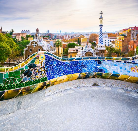 Colorful mosaic bench and cityscape view of Barcelona from Park Güell, featuring ornate architecture and trees under a partly cloudy sky.