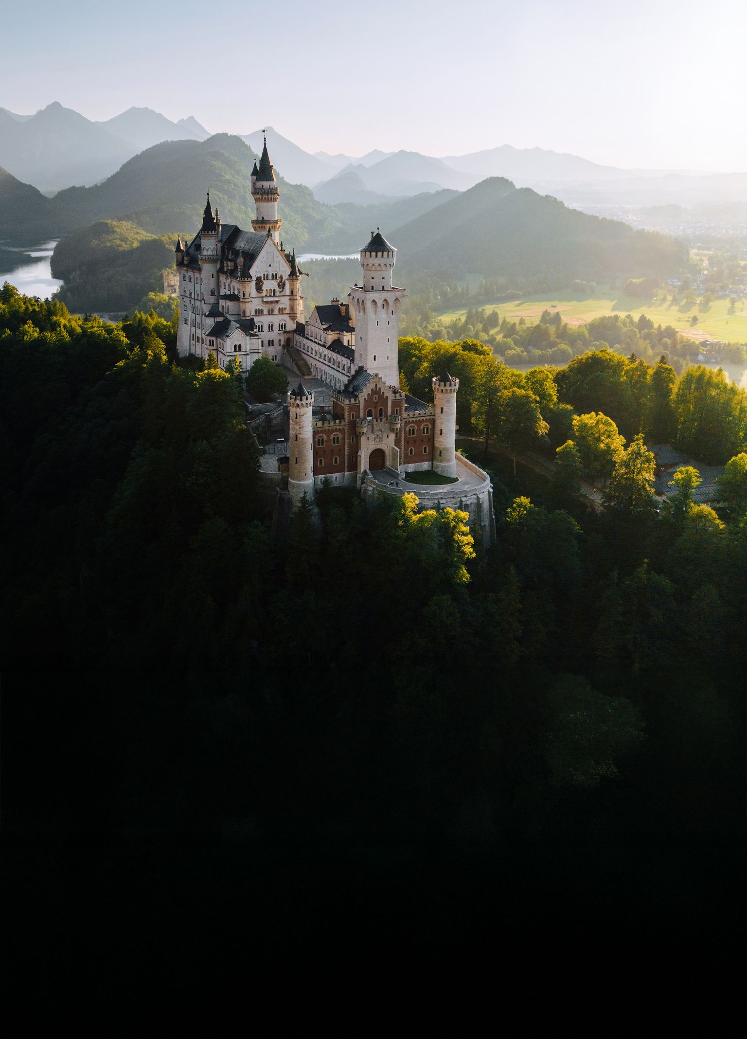 A fairytale castle surrounded by lush forests, with a mountainous landscape in the background under a clear sky.