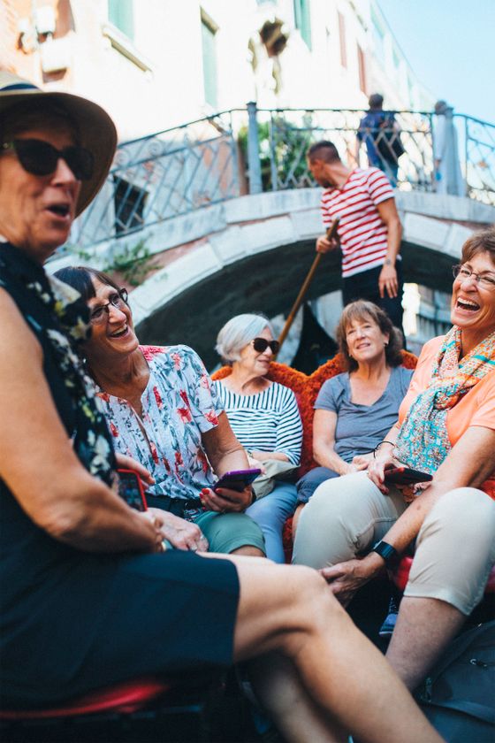 Group of women laughing and sitting in a gondola in Venice, with a gondolier standing behind them near a stone bridge.