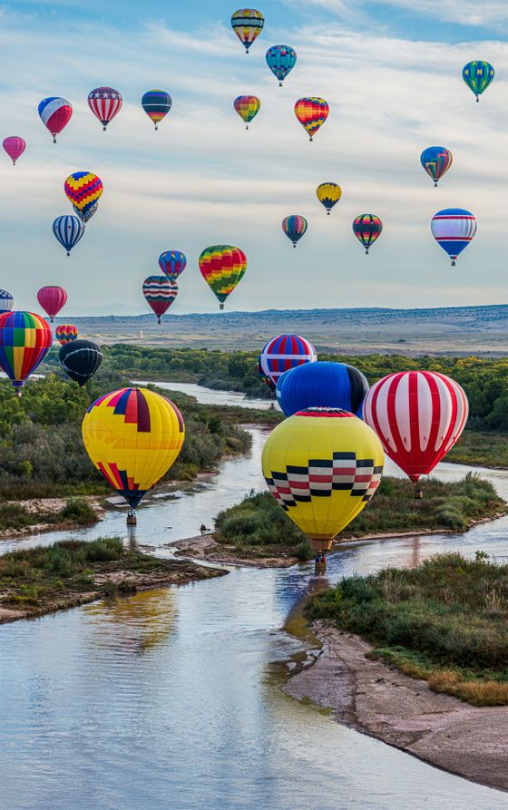 Colorful hot air balloons float above a winding river and lush landscape under a partly cloudy sky.