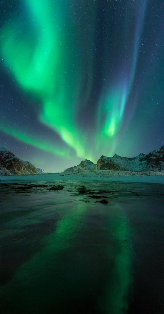 Vibrant green Northern Lights illuminate a snowy mountain landscape, reflecting on a dark and tranquil body of water.