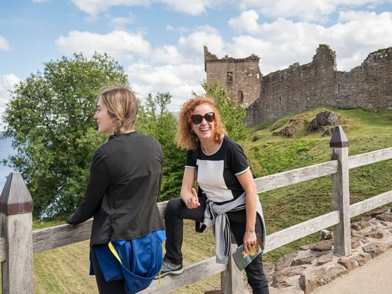 Two women lean against a wooden fence with stone ruins in the background and a lake to the left. The woman on the right wears sunglasses and gestures with her finger, while the woman on the left looks toward the water.