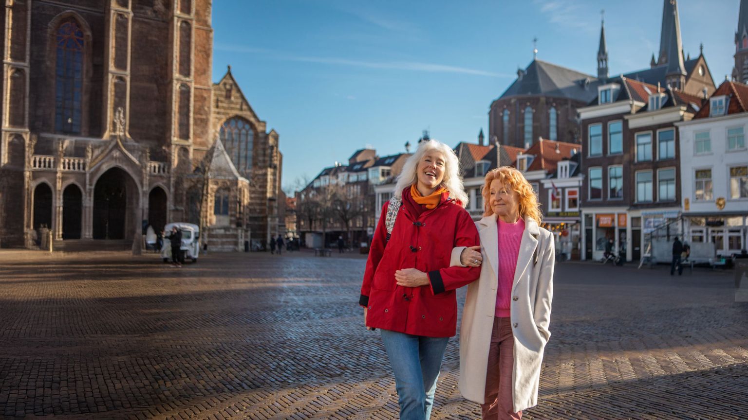 Two women walking arm in arm in a sunny cobblestone European square, with historic buildings in the background.