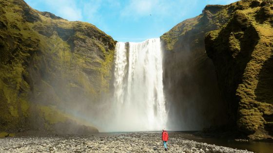Person in a red jacket stands near a large waterfall cascading between mossy cliffs, under a bright blue sky. A faint rainbow is visible.