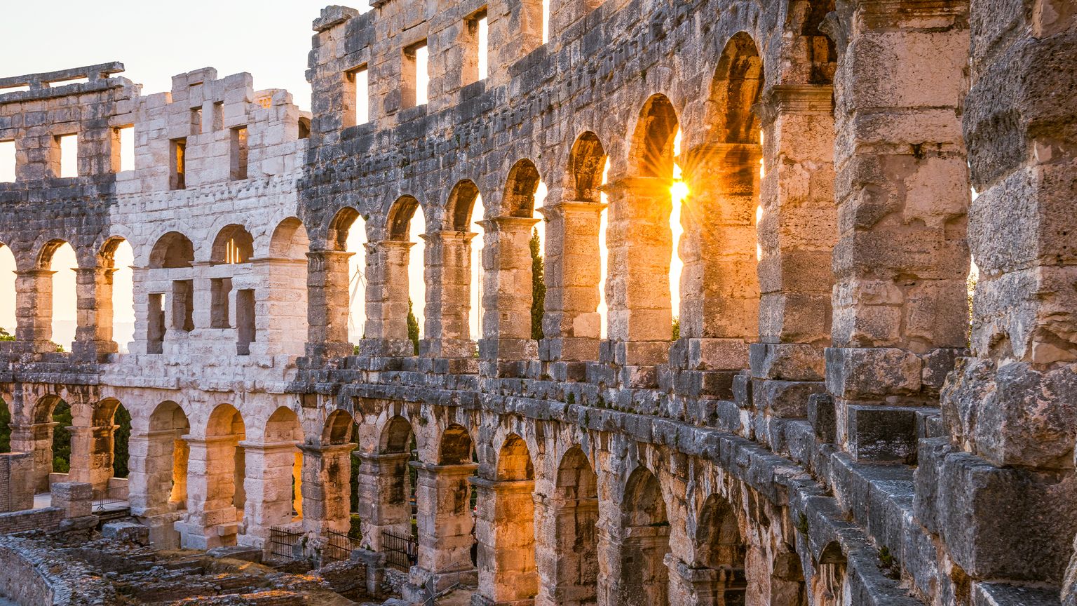 Historic stone amphitheater with multiple arches, illuminated by the setting sun peeking through, casting a warm glow.