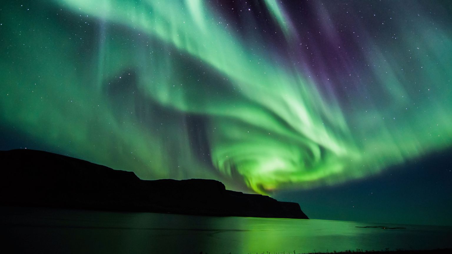 Green and purple aurora lights swirl in the night sky over dark hills and a calm water surface.
