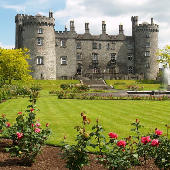 Kilkenny Castle with grey stone walls and circular towers stands behind a manicured green lawn with flower beds and a spraying fountain. Large trees flank both sides of the castle, and stone steps lead up to the central entrance.