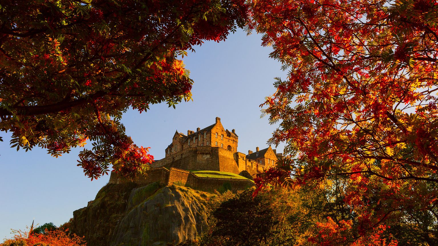 A castle sits atop a rocky hill, framed by vibrant autumn foliage under a clear blue sky.