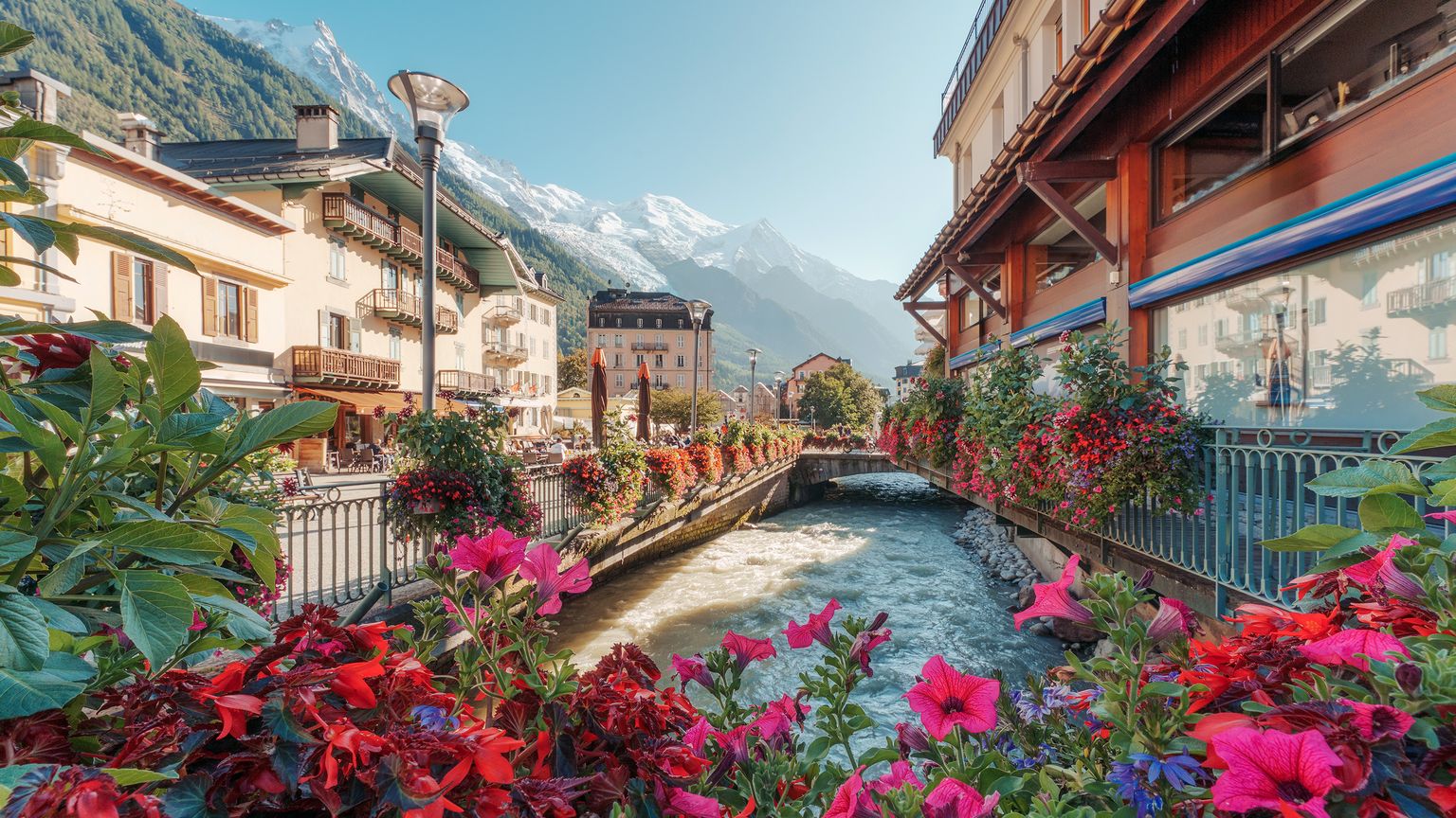 A scenic view of a canal lined with vibrant flowers, surrounded by alpine-style buildings and snow-capped mountains in the background.