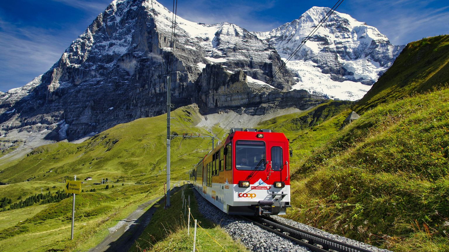 Red train traveling on a mountain railway with stunning snow-capped peaks in the background and lush green hills on either side.