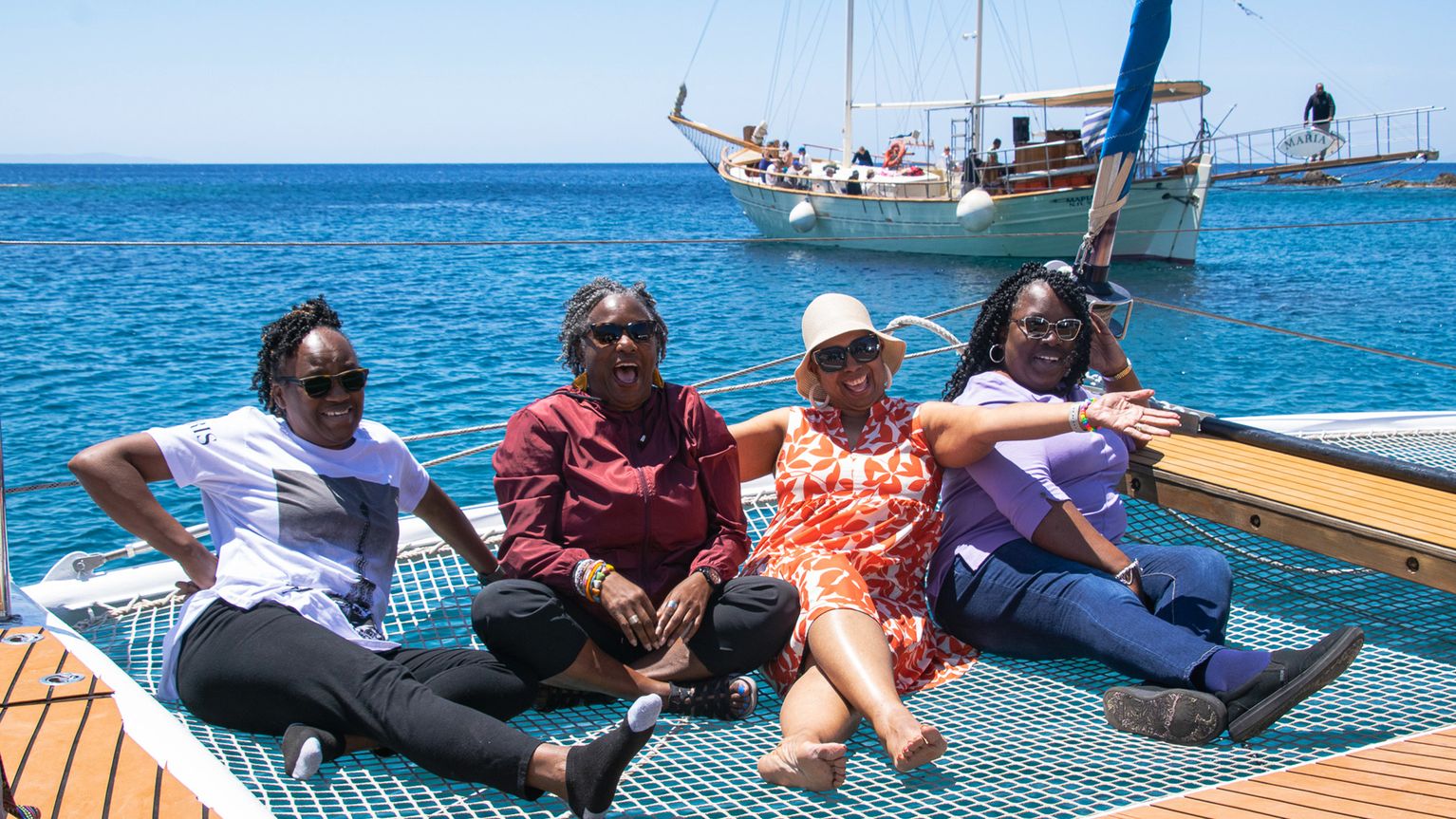 Four women sitting on a boat net, smiling and relaxing by the sea with a sailboat in the background.