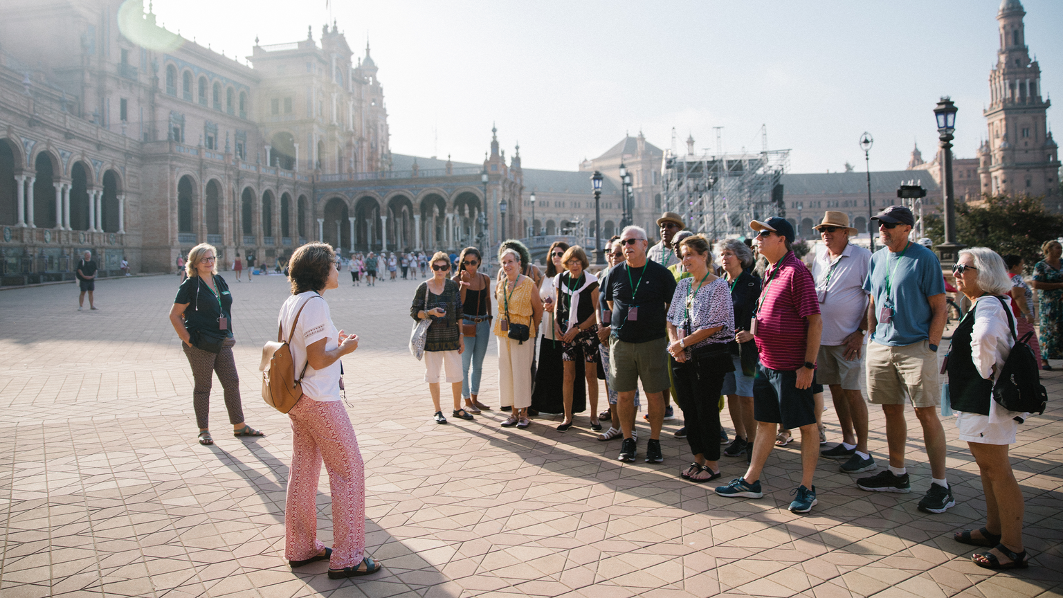A tour guide addresses a group of tourists in a sunlit plaza with historic architecture, featuring archways and towers in the background.