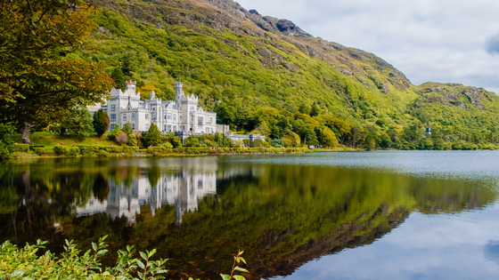 Kylemore Abbey, a stone castle with towers, sits on the edge of a lake at the base of a large, green, tree-covered mountain. The architecture of the abbey is reflected clearly in the calm water of the lake.