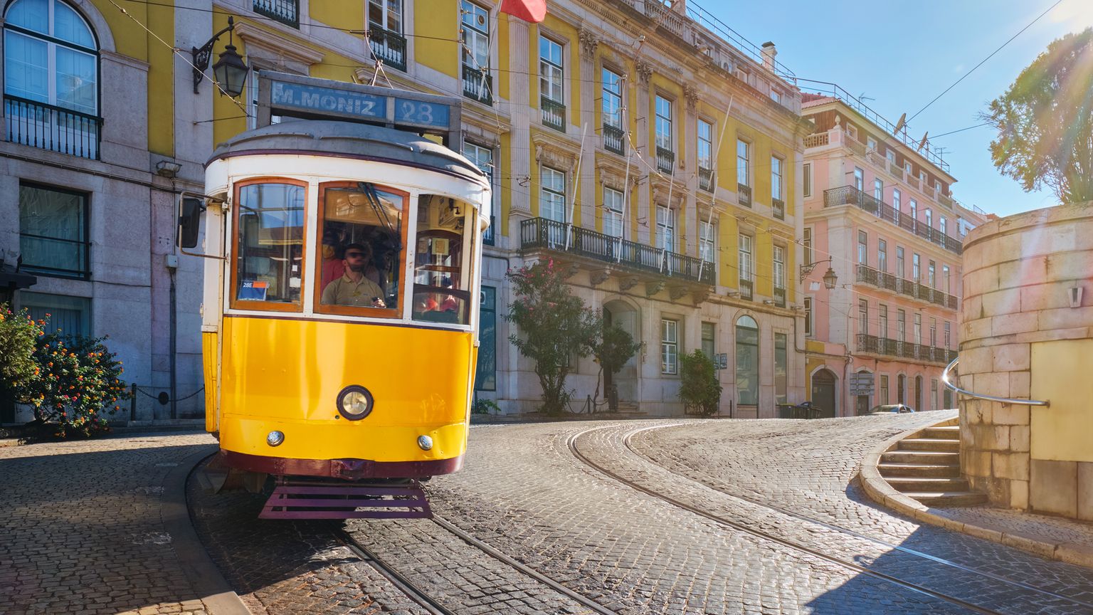 A vibrant yellow tram travels on a cobblestone street in a sunny European city, lined with historic buildings.