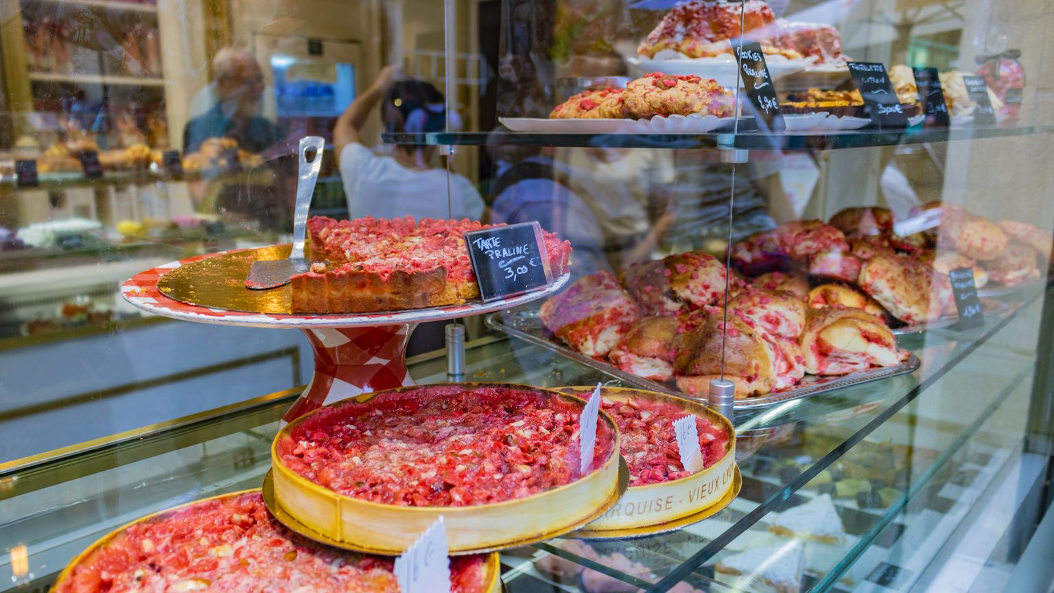 Bakery display with praline tart, cookies, and pastries on glass shelves, labeled with prices. People can be seen through the glass.