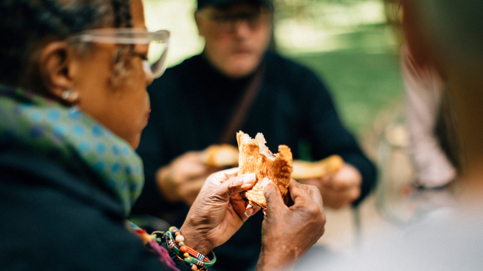 A woman holds a piece of bread, with people in the background, sitting outdoors in a park setting.