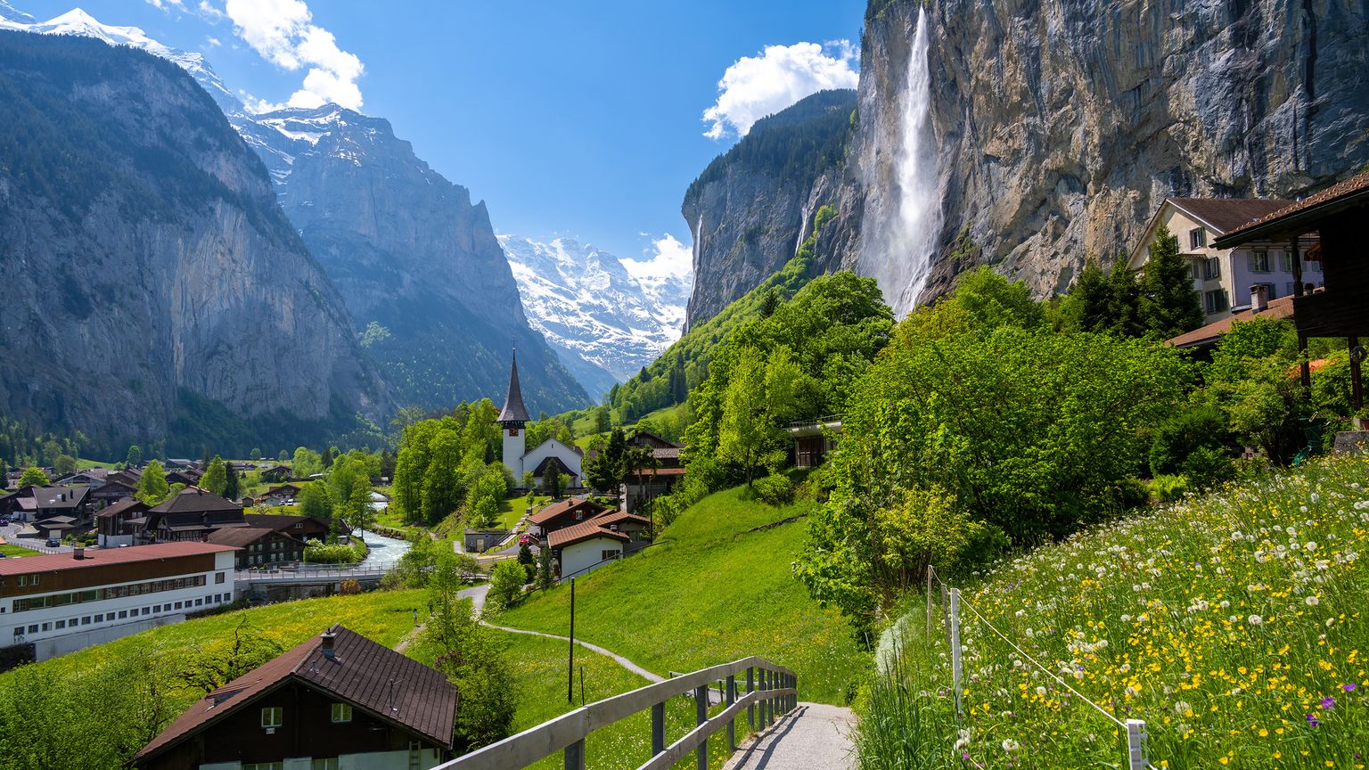 Mountain village with a church, waterfall, and wooden houses set against towering cliffs and snow-capped peaks under a bright blue sky.