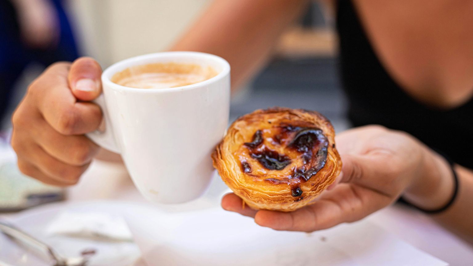 Person holding a cappuccino in a white mug and a pastel de nata, a Portuguese custard tart, on a table.