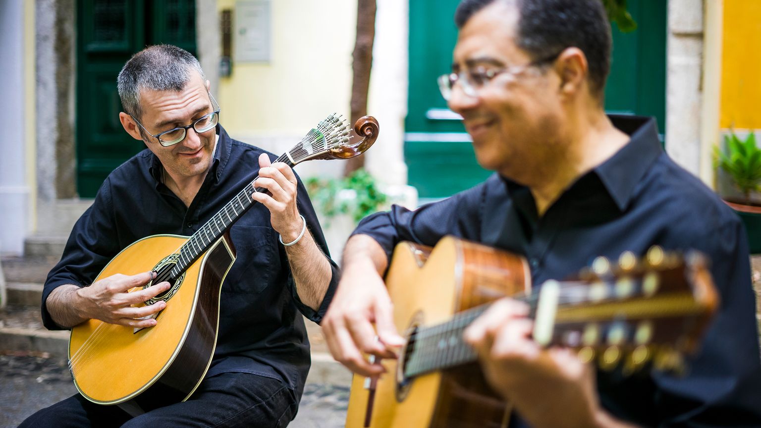 Two musicians playing string instruments outdoors; one with a mandolin, the other with a guitar. Both wear black shirts and seem focused and content.