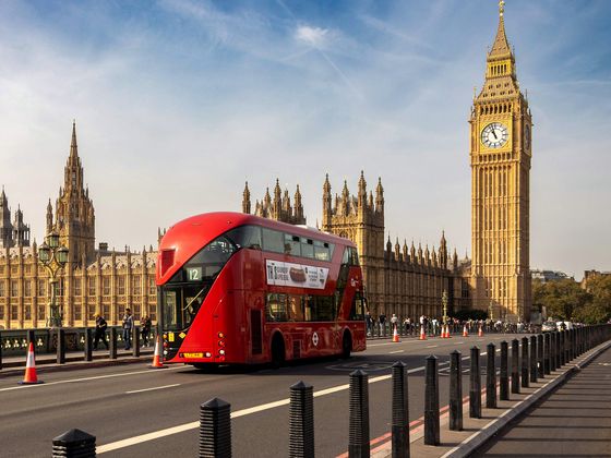 A red double-decker bus drives on Westminster Bridge with the Houses of Parliament and Big Ben in the background under a clear blue sky.