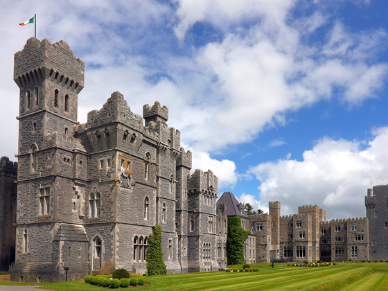Ashford Castle, a stone structure with multiple towers and turrets, sits on a grassy lawn under a blue sky with white clouds, with an Irish flag flying from the highest tower.