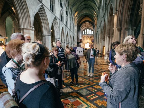 A tour guide speaks into a small device while leading a group of people inside a cathedral with vaulted ceilings and patterned tile floors. The attendees are gathered around the guide, many looking toward the cathedral's interior.
