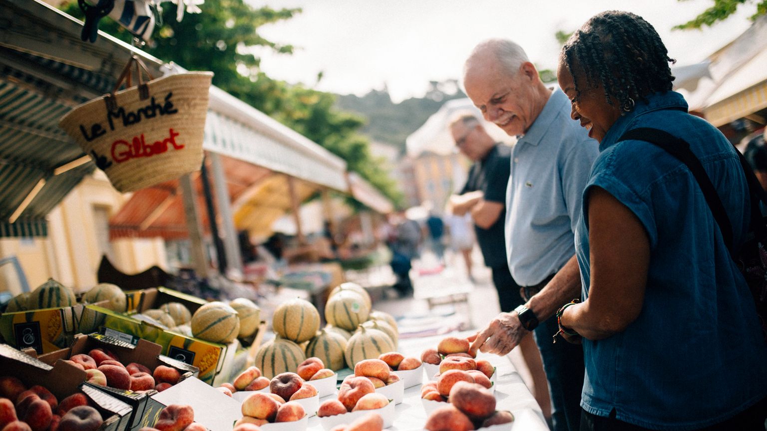 Two people browsing fresh produce at an outdoor market stall on a sunny day, with signs and melons in the background.