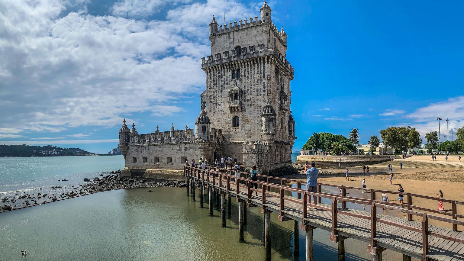 Belem Tower in Lisbon, Portugal, stands surrounded by water, with a wooden walkway leading to it, against a partly cloudy blue sky.