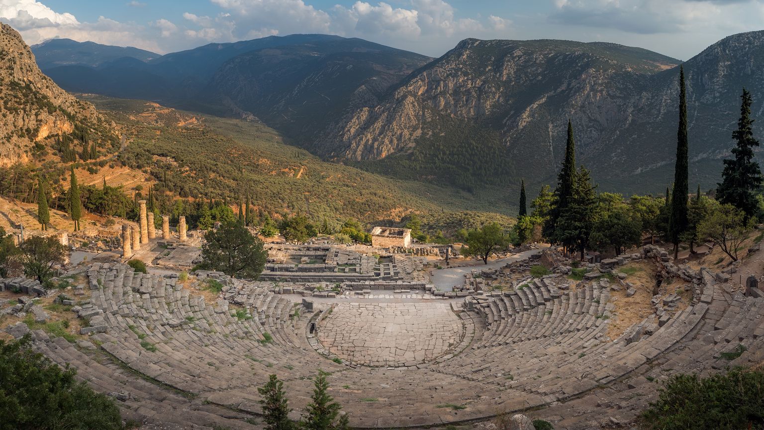 Ancient stone amphitheater in a mountainous landscape, with scattered ruins and cypress trees under a partly cloudy sky.