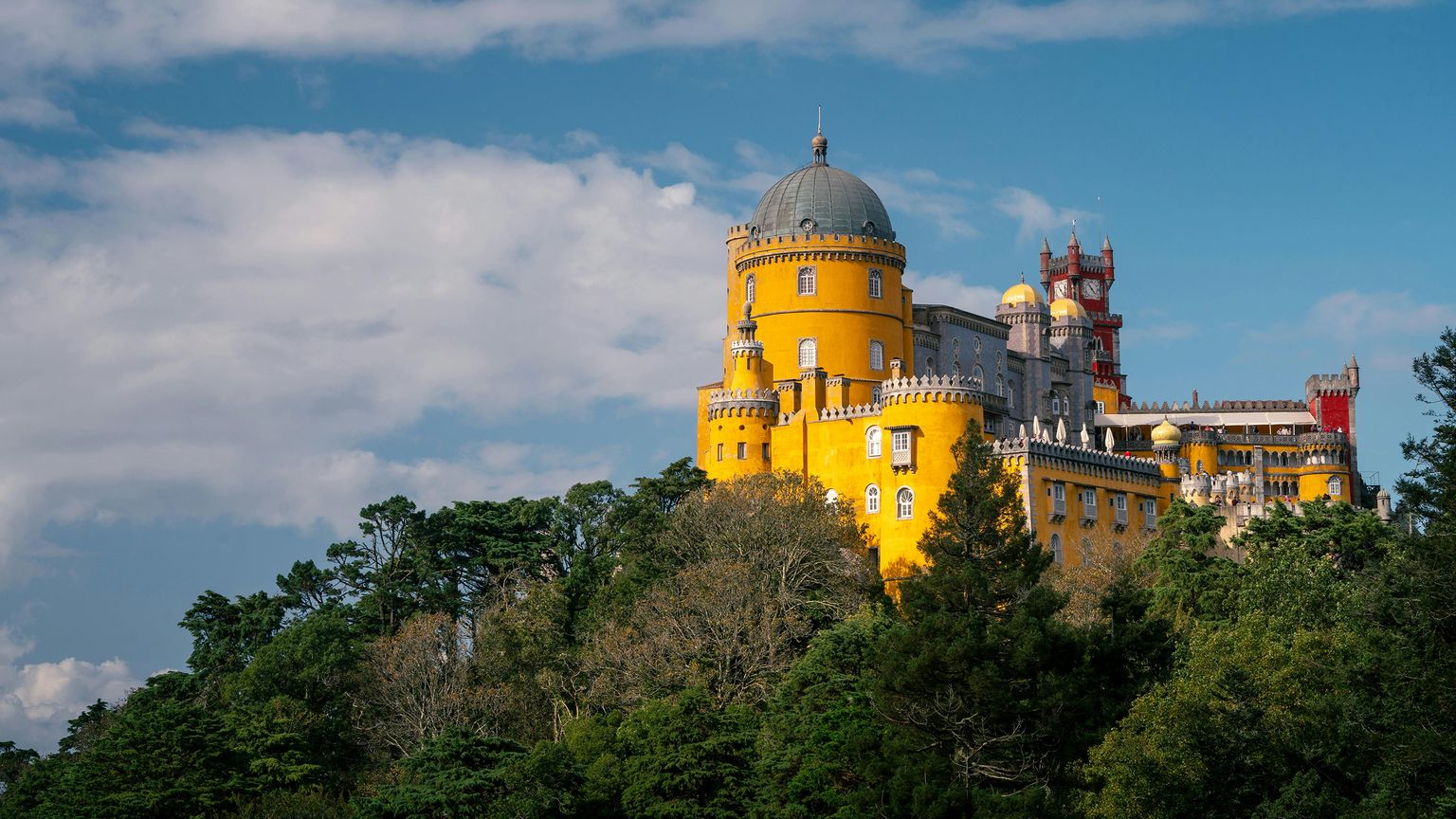 Bright yellow, multi-turreted castle with a red spire atop a lush, tree-covered hill under a blue, partly cloudy sky.
