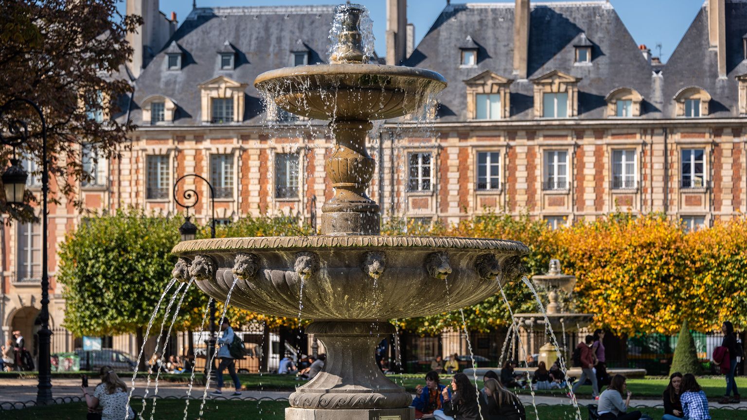 A fountain with decorative features and flowing water in a vibrant park, surrounded by historic buildings, trees, and people relaxing in the sun.