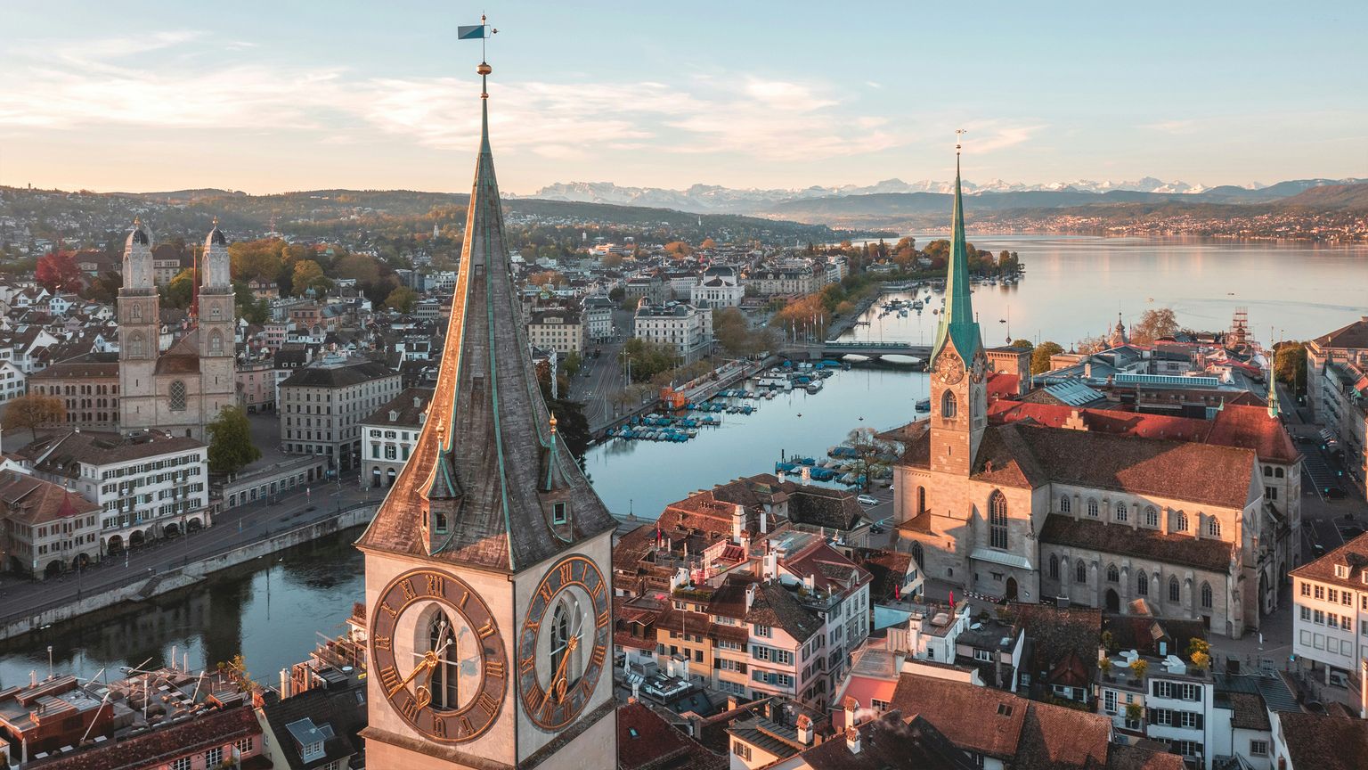 Aerial view of Zurich at sunset, featuring church towers, historic buildings, a serene river with boats, and mountains in the background.
