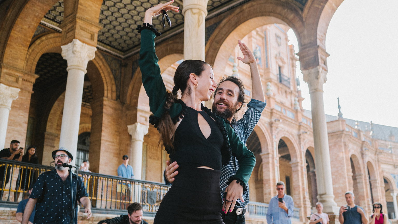 A couple dances passionately under a historic archway, surrounded by onlookers. The scene captures a lively outdoor performance.