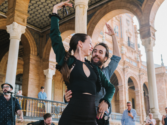 A couple dances passionately under a historic archway, surrounded by onlookers. The scene captures a lively outdoor performance.