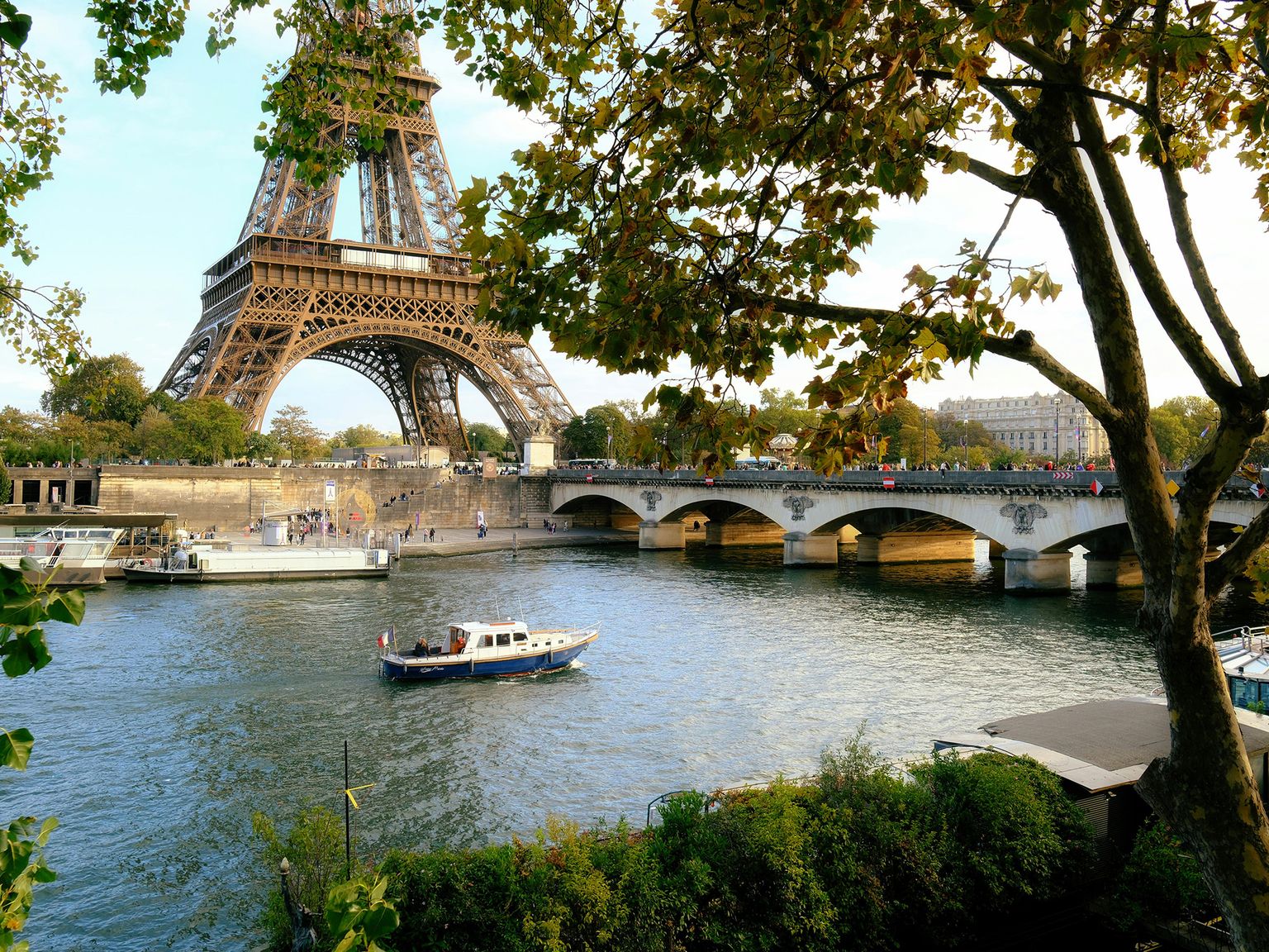 Eiffel Tower with a bridge over the Seine River in Paris, surrounded by trees and boats on the water.