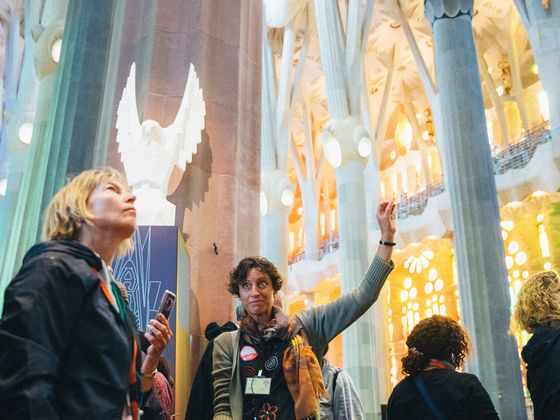 A group of tourists inside a cathedral admire the architecture, with a guide pointing upward, surrounded by vibrant stained glass light.