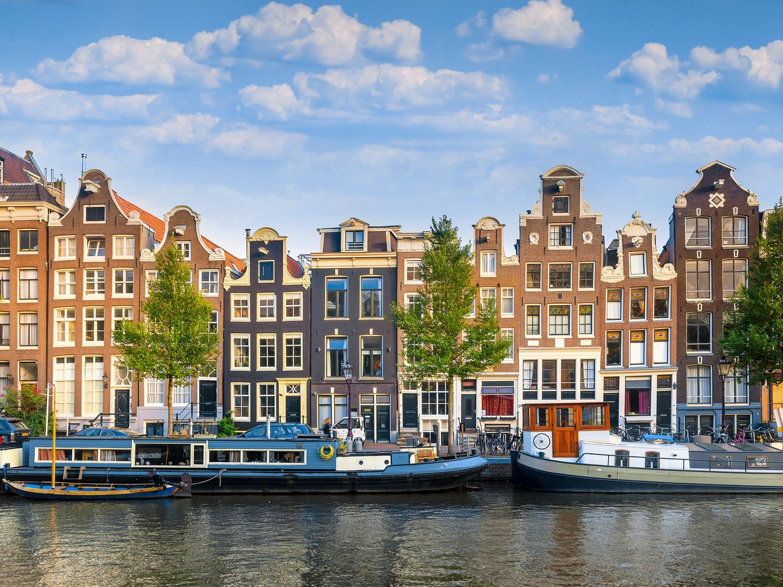Canal in Amsterdam with traditional Dutch houses and moored boats under a blue sky with clouds.