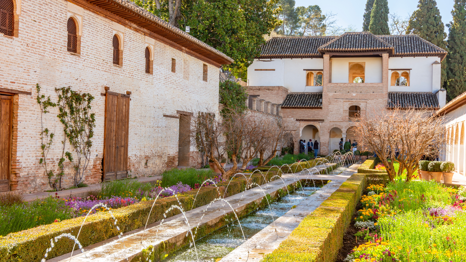 A picturesque garden with a long fountain, surrounded by blooming flowers and hedges, leads to a historic, white-brick building under a clear sky.