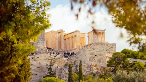 View of the Acropolis in Athens, Greece, framed by lush trees, with tourists exploring the ancient ruins under a clear sky.