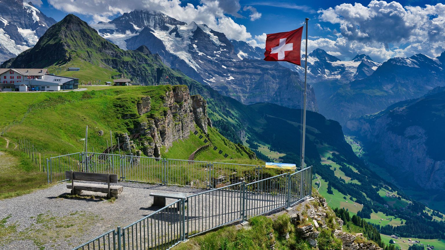 Scenic view of Swiss Alps with snow-capped peaks, green valleys, a Swiss flag, and a bench in the foreground under a cloudy sky.