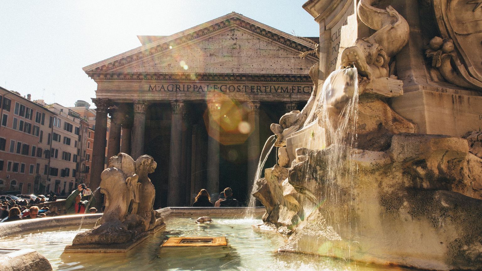 A sunlit scene of the Pantheon in Rome, with a decorative stone fountain in the foreground, and people gathered around.