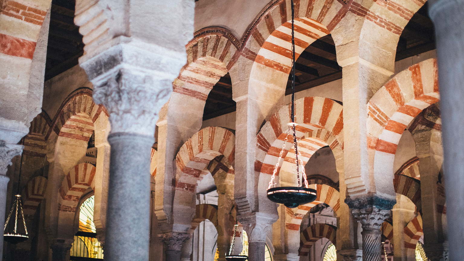 Interior of the Mosque-Cathedral of Córdoba, featuring iconic red and white striped arches and ornate columns. Hanging lamps add soft lighting.
