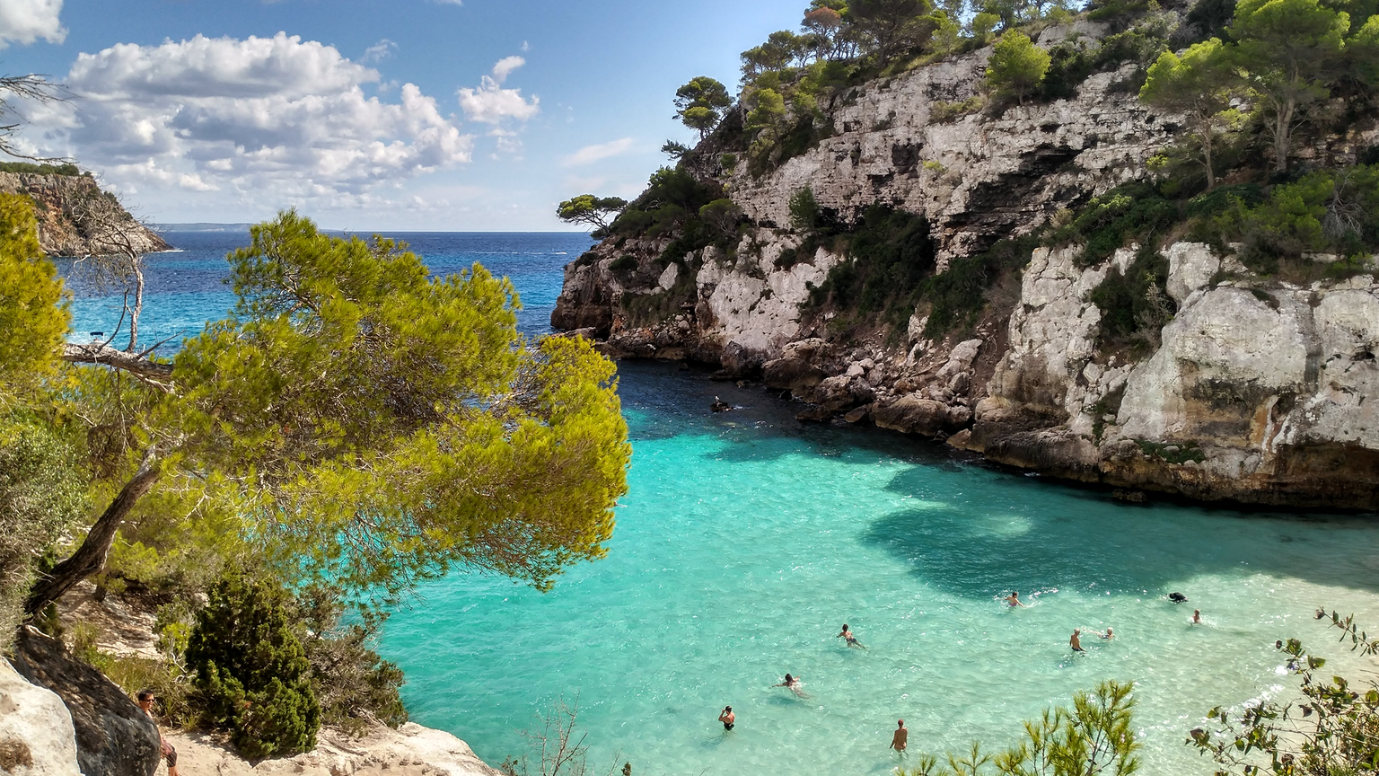 A scenic view of a rocky coastal cove with turquoise waters, surrounded by cliffs and trees, where people are swimming under a partly cloudy sky.