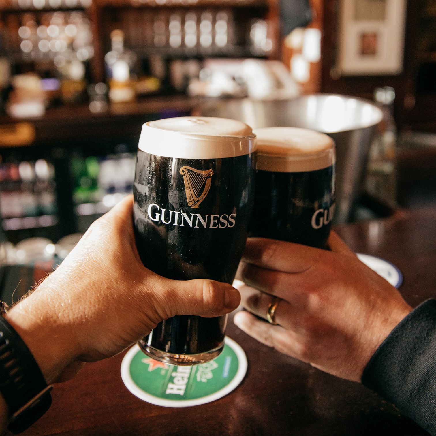 Two hands holding pints of Guinness at a bar, with a background of bottles and a wooden counter.