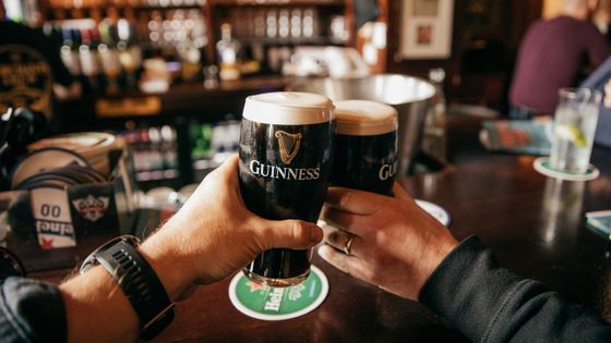Two hands holding pints of Guinness at a bar, with a background of bottles and a wooden counter.