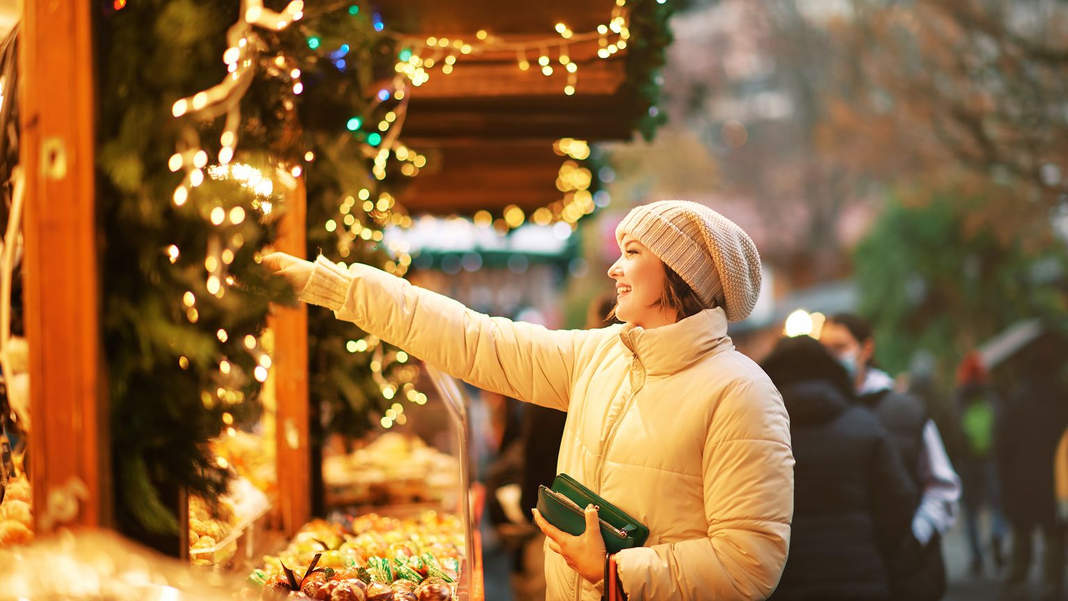 A woman in a beige coat and knitted hat smiles while reaching for items at a festive market stall adorned with string lights and holiday decorations.