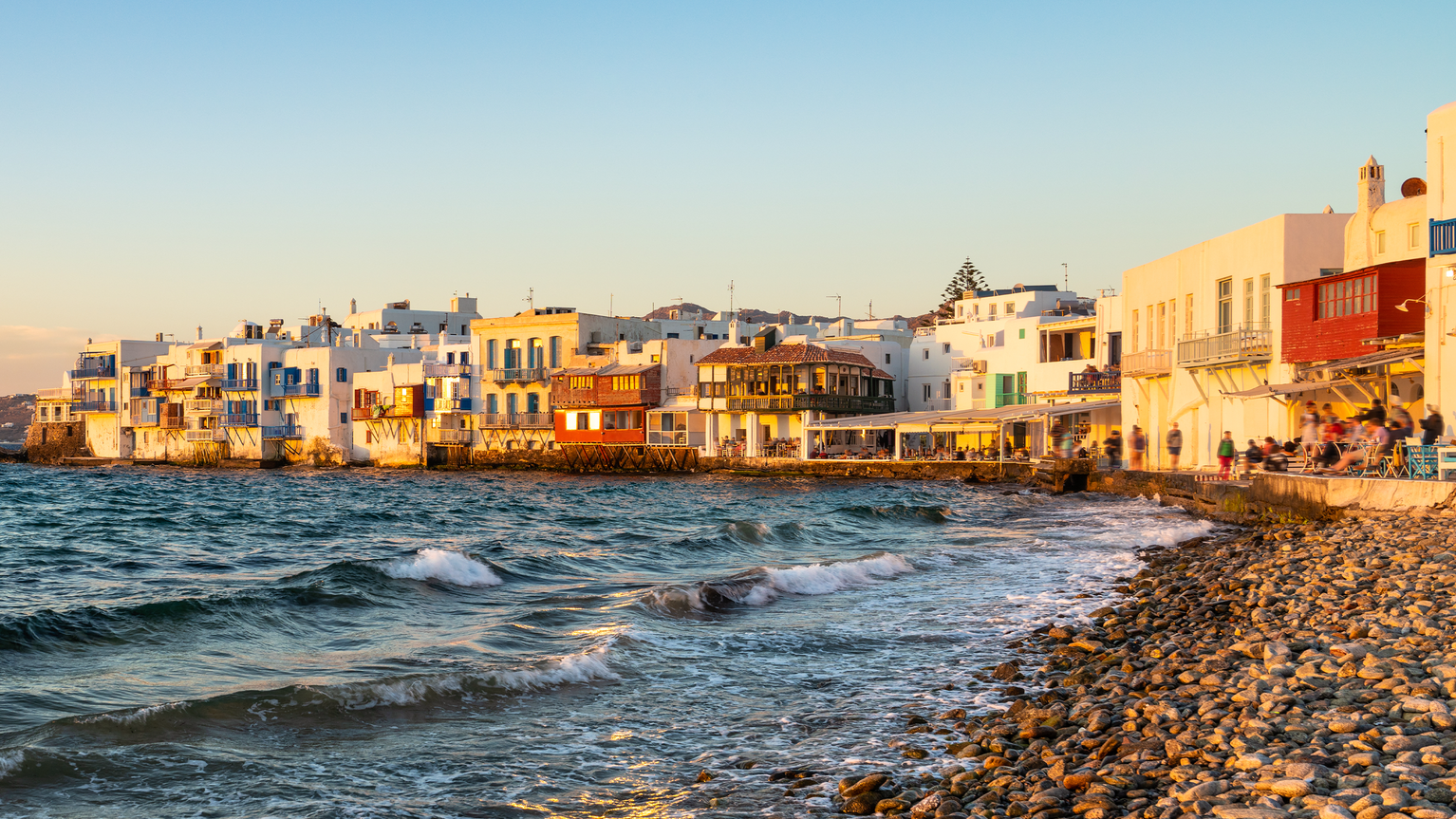 Sunset view of white buildings along a rocky shoreline in Mykonos, Greece, with waves gently crashing onto the beach.