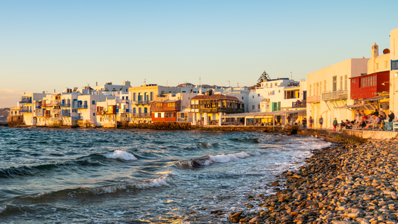 Sunset view of white buildings along a rocky shoreline in Mykonos, Greece, with waves gently crashing onto the beach.